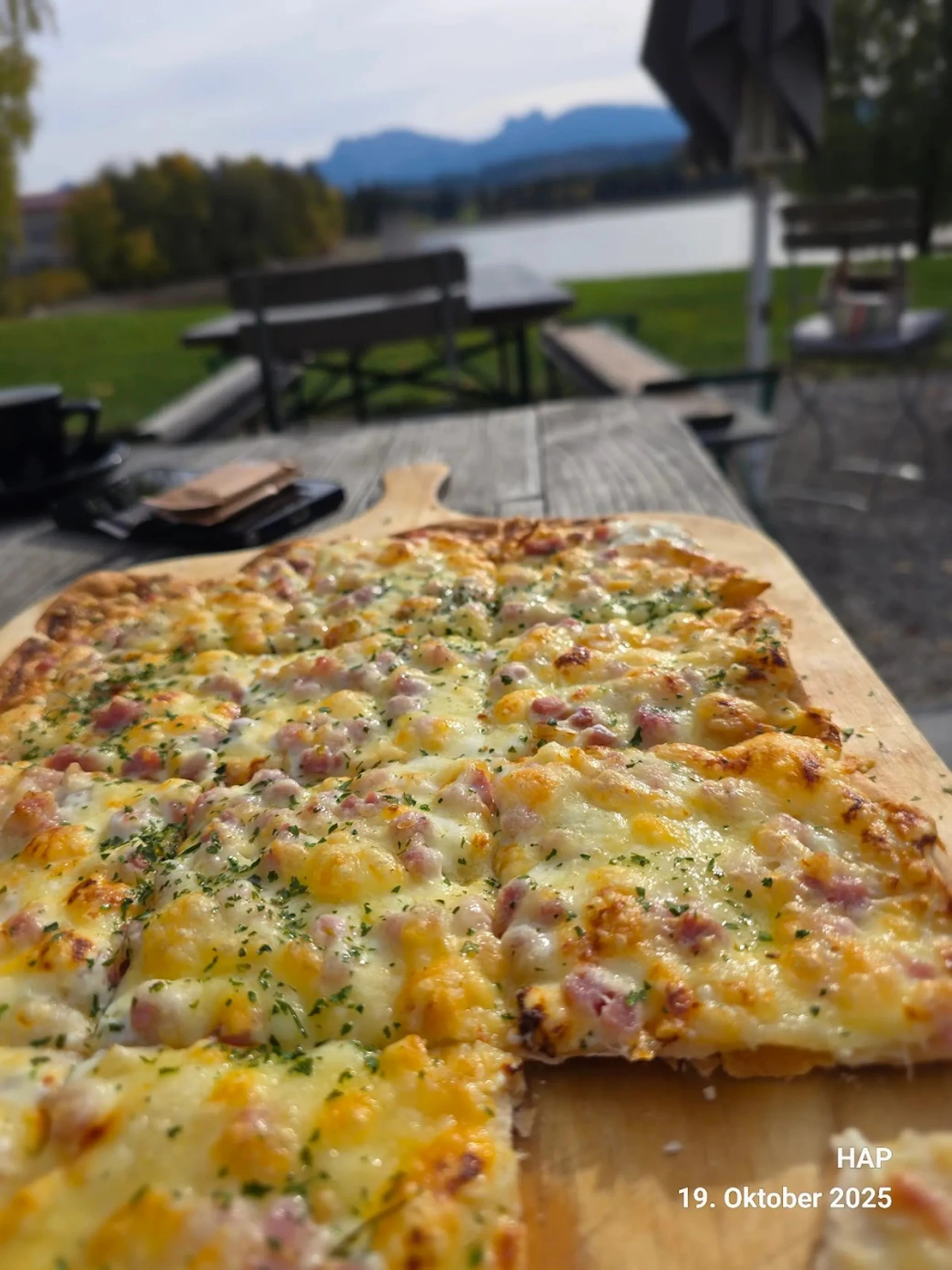 Eine Käse-Schinken-Pizza auf einem Holzbrett mit Blick auf einen See und Berge.