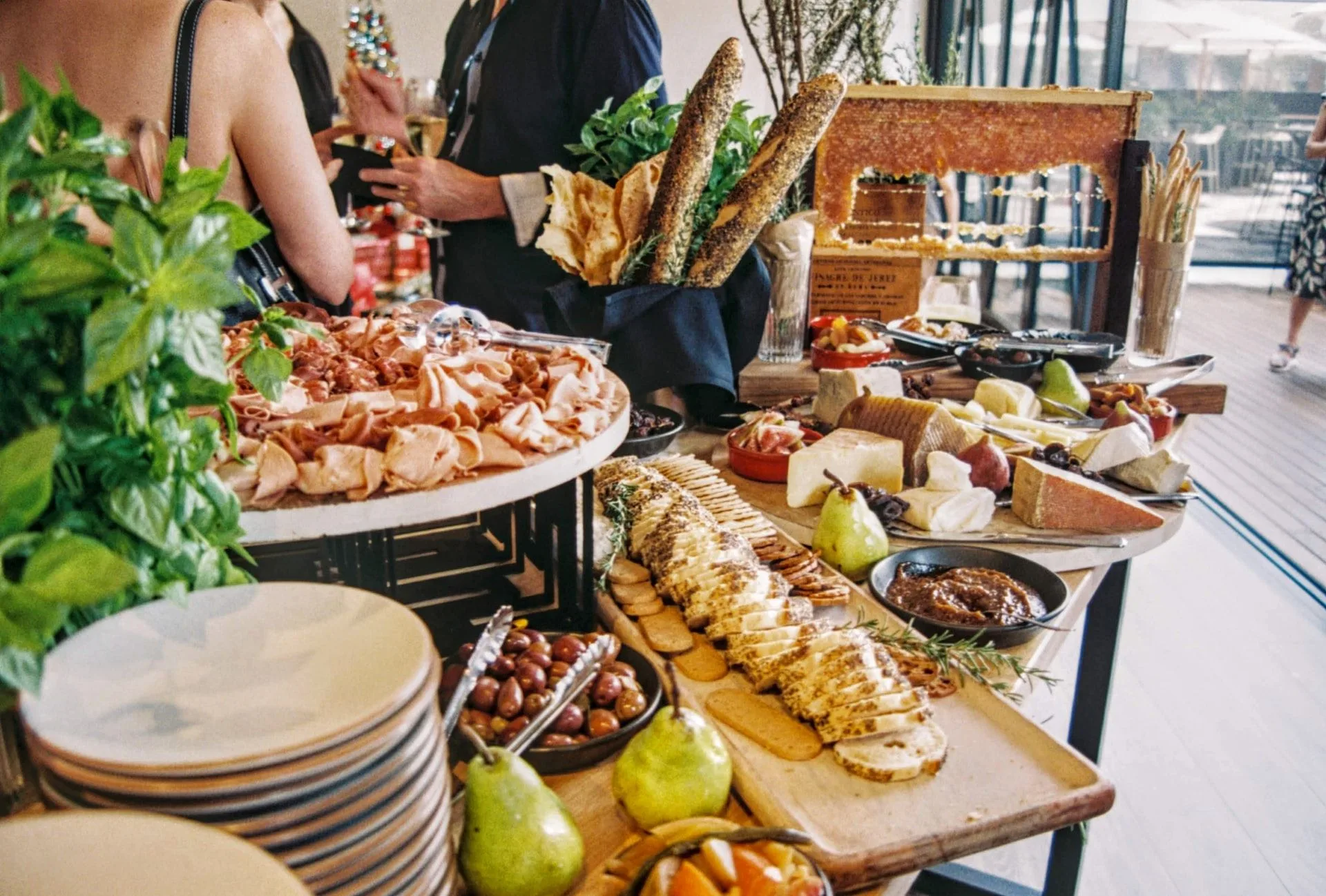 Großes Buffet mit Aufschnitt, Käse, Brot, Oliven, Birnen und Kräutern, mit Menschen im Hintergrund.